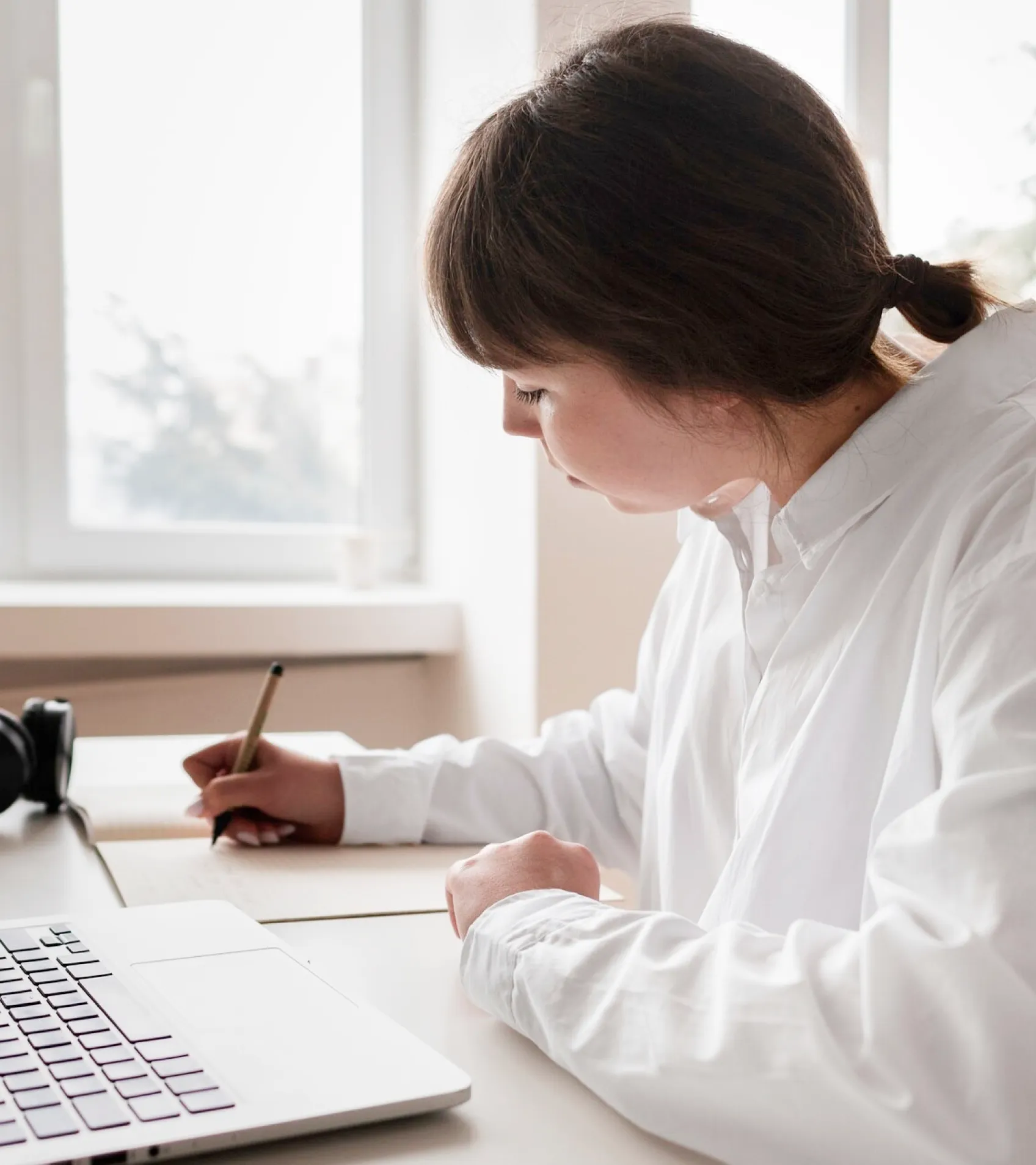 A woman dressed in a white shirt is engaged in writing on her laptop, illustrating a scene of concentration and creativity.